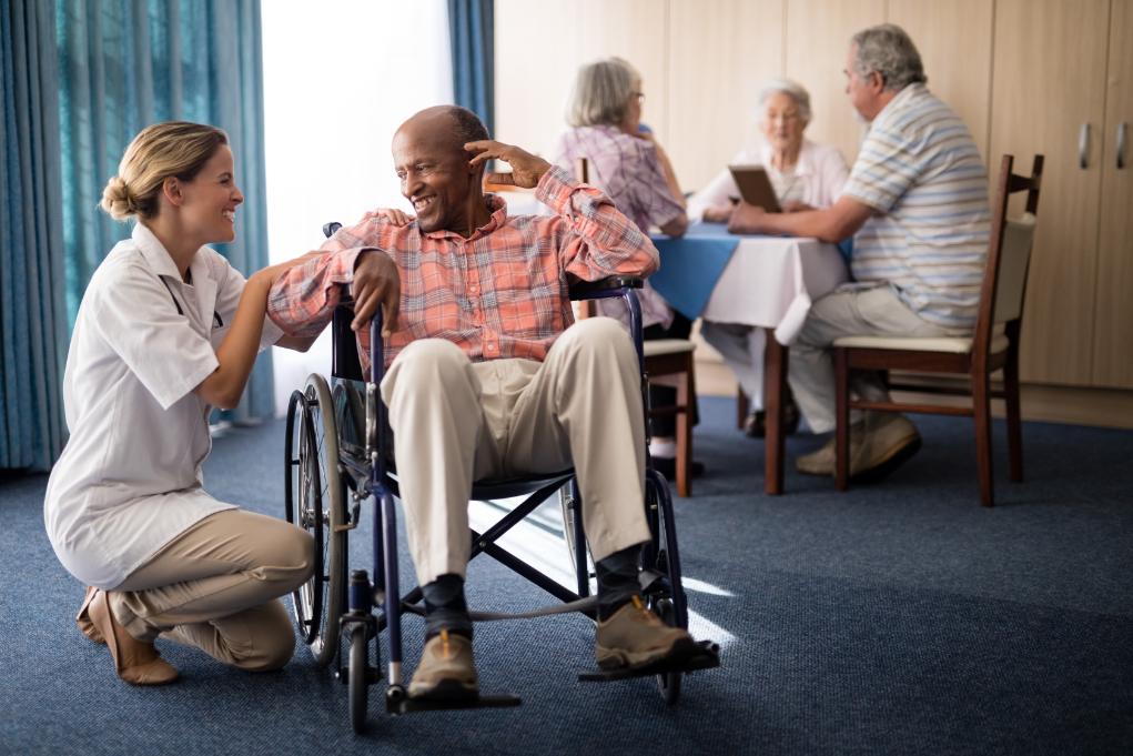 old man sitting on a wheel chair assisted by a female nurse and a group of seniors at the back of the room